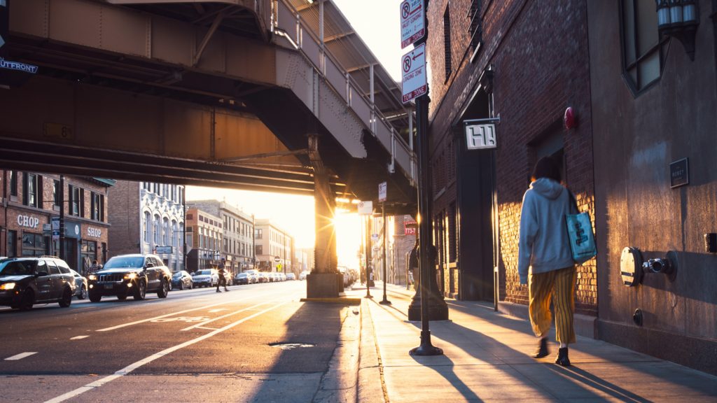 A woman walking down a city street with the sun shining between buildings
